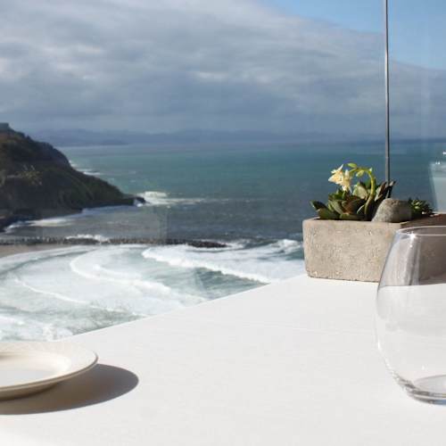 Ambiente de comedor sereno con vista al océano, decorado con una planta pequeña, ideal para una experiencia gastronómica tranquila y elegante.