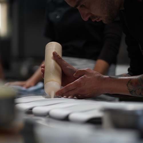 Chef aplicando una crema suave con una manga pastelera, en un entorno de cocina bulliciosa.