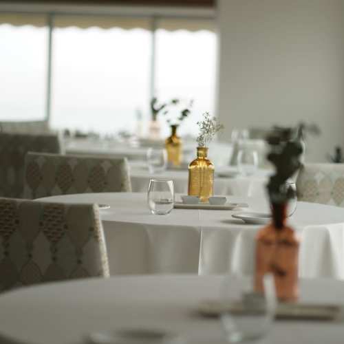 Interior de restaurante moderno con mesas elegantemente vestidas, decoradas con centros de mesa de jarrones delgados y vegetación delicada, creando un ambiente acogedor y minimalista.