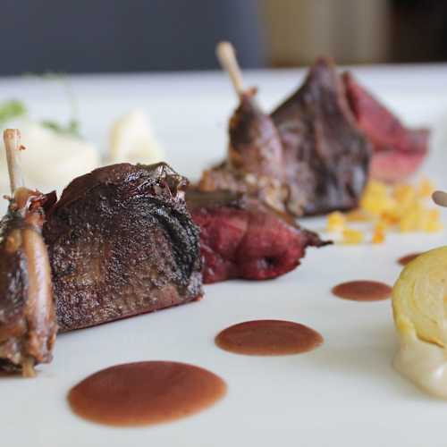 Piezas tiernas de carne de caza con salsa brillante, acompañadas de puré cremoso y rodajas de un vegetal amarillo, decoradas con microgreens y hierbas.