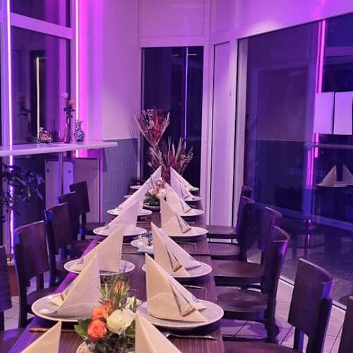 Intérieur de restaurant moderne avec une grande table élégamment dressée, ornée de petites compositions florales et d'un éclairage ambiant violet, créant une atmosphère accueillante.