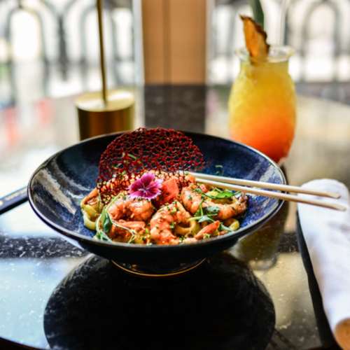 Camarões suculentos cobertos com um molho rico de sabores doces e salgados, guarnecidos com microverdes e um crocante vermelho, sobre fatias finas de noodles salteados. Acompanha um cocktail colorido com camadas de laranja e amarelo, decorado com uma fatia de ananás.