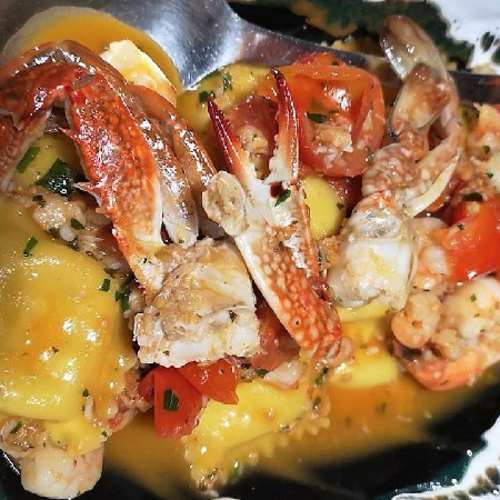 Ravioli di mare ripieni di crema, guarniti con granchio fresco e gamberi, serviti in una salsa leggera e aromatica con pomodori e erbe tritate.