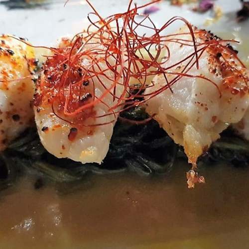 Tre pezzi di frutti di mare delicatamente cotti, probabilmente capesante, serviti su un letto di verdure saltate, con una salsa cremosa e un tocco di peperoncino rosso o zafferano.