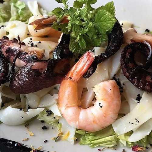 Piatto di frutti di mare con gamberi e polpo su un letto di fettuccine, guarnito con menta e condimento nero, servito su un letto di lattuga croccante.