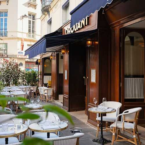 Extérieur d'un restaurant charmant avec de grandes fenêtres élégantes, des tables bien disposées avec nappes blanches, et une ambiance accueillante agrémentée de verdure.