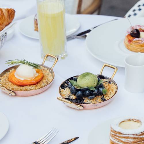Sélection de délicieuses pâtisseries et desserts pour le petit-déjeuner, comprenant un œuf poché crémeux sur un lit de grains dorés, un mélange de myrtilles avec une boule de glace au thé vert, un croissant doré, une tarte aux fraises, et une pâtisserie délicate. Accompagné de jus d'orange et d'une mimosa, ainsi qu'une tasse de thé.