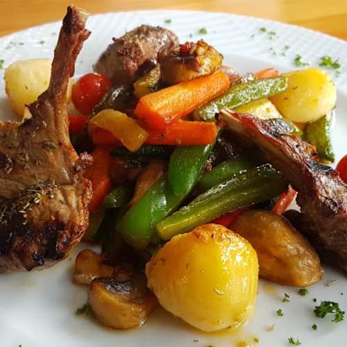 Gegrilde lamskoteletten met kleurrijke gebakken groenten zoals groene paprika, wortelen en cherrytomaten, vergezeld van goudbruine baby-aardappelen en gegarneerd met fijngehakte kruiden.