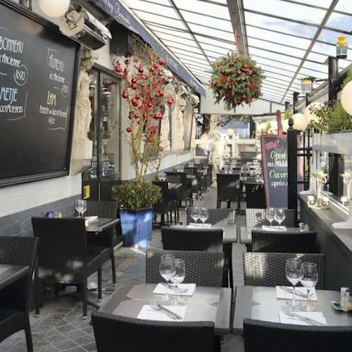 Het interieur van het restaurant is charmant en luchtig, met weelderige planten en decoratieve bloemstukken die een uitnodigende sfeer creëren. De tafels zijn netjes gedekt en de zitplaatsen zijn comfortabel met donkere rieten stoelen. Grote ramen laten natuurlijk licht binnen, wat de uitnodigende uitstraling versterkt. Op de muren zijn krijtbordmenu's te zien, wat een rustieke toets aan de inrichting toevoegt. De indeling suggereert een gezellige en elegante eetervaring.