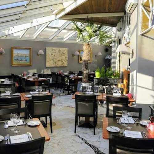 Heldere en luchtige restaurantinterieur met een glazen dak, potted planten en kunst aan de muren, wat bijdraagt aan de sfeer. De indeling biedt een comfortabele eetervaring met zowel intieme zitplaatsen als ruimte voor grotere groepen.