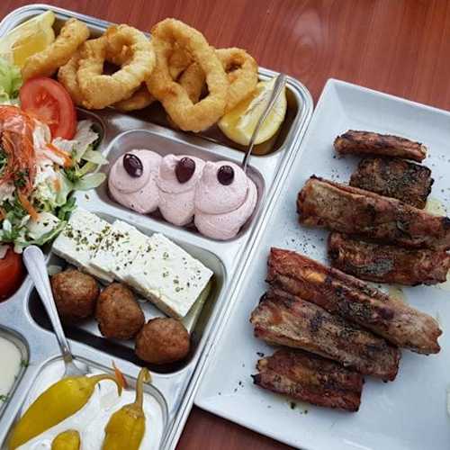 Groot plateau met diverse gerechten, waaronder krokante uienringen, een frisse salade met tomaten, sla en garnalen, romige mousse en witte kaas. Daarnaast gegrilde spareribs met een rokerige smaak, coleslaw en gouden frietjes, vergezeld van schijfjes citroen.