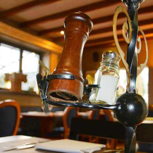 Rustieke pepermolen van donker hout en glazen zoutvaatje, gepresenteerd op een metalen houder, in een gezellige restaurantinterieur met houten balken en sfeervolle verlichting.