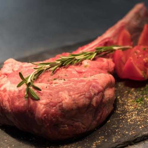 Een grote, rauwe tomahawk steak met indrukwekkende marmering en rijke rode kleur, vergezeld van twee in bloemblaadjes gesneden tomaatpartjes en een takje rozemarijn. Licht gekruid, klaar voor het grillen of roosteren.