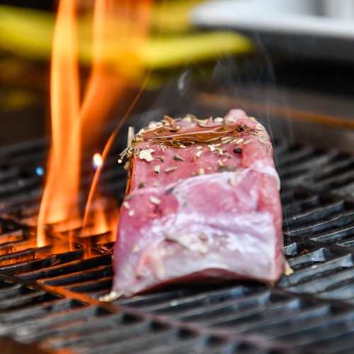 Un morceau de viande, probablement un steak, grillé sur une flamme vive, avec une couleur rougeâtre indiquant une cuisson à point. Des herbes, peut-être du romarin ou du thym, libèrent des huiles aromatiques. Les marques de grillade suggèrent une croûte bien saisie, tandis que les flammes ajoutent une intensité et une saveur fumée au processus de cuisson.