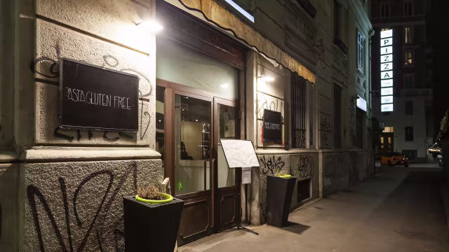 Ristorante con estetica moderna e urbana, ingresso con porte in vetro e struttura in legno, illuminato da luci soffuse e piante alte ai lati. Insegna con il nome del ristorante e focus su opzioni senza glutine.