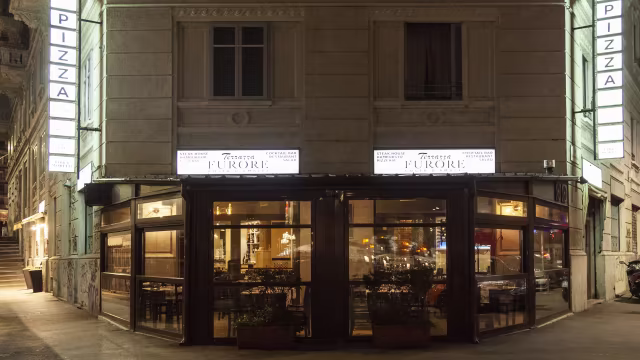 Ristorante accogliente con grandi finestre, specializzato in pizza, illuminato dolcemente e con piante in vaso all'ingresso.