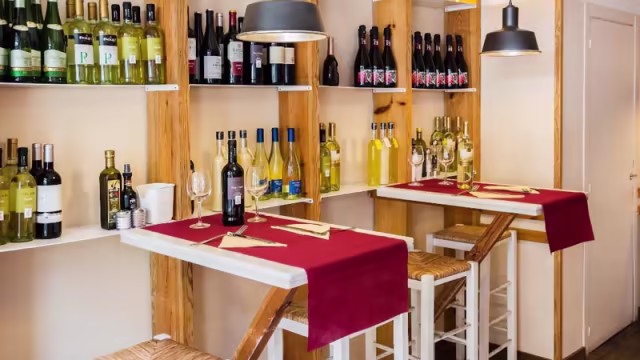 Interior acogedor de un restaurante con estanterías de madera llenas de botellas de vino, mesas altas con manteles rojos y una iluminación suave que crea un ambiente íntimo.