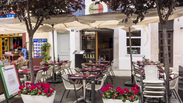 Área de comedor al aire libre con mesas bajo sombrillas, sillas blancas y manteles oscuros, rodeada de vegetación y flores rosas.