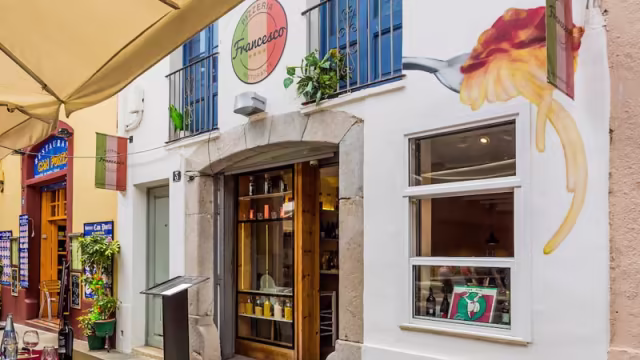 Restaurante acogedor con un mural de un tenedor y pasta, entrada con puerta de madera y grandes ventanas, rodeado de plantas en macetas, ideal para una experiencia gastronómica italiana.