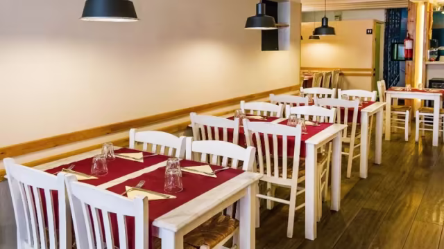 Interior de un restaurante acogedor con mesas y sillas de madera blanca, manteles rojos y una atmósfera cálida iluminada por luces colgantes.