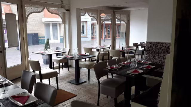 Het restaurantinterieur heeft een modern ontwerp met een mix van lichte en donkere tinten. De tafels zijn netjes gerangschikt met witte borden en rode servetten, wat een opvallend contrast creëert. De comfortabele beige stoelen en de donkere houten tafels zorgen voor een verfijnde uitstraling. Grote ramen laten natuurlijk licht binnen, wat de uitnodigende sfeer versterkt. Decoratieve elementen, zoals patroonwanden en subtiele groenvoorzieningen, voegen warmte toe aan de algehele ambiance.