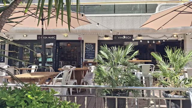 Área de comedor al aire libre con mesas de madera, sillas de metal blanco y plantas en macetas, creando un ambiente acogedor y moderno.