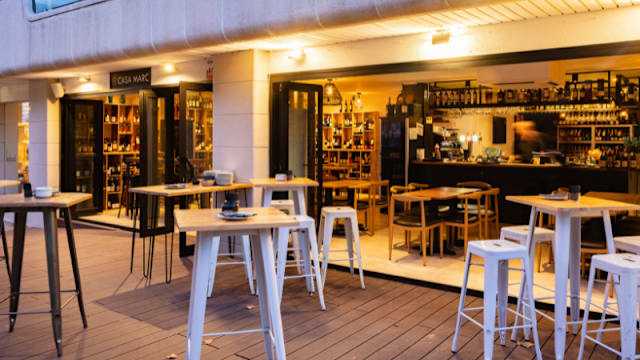 Restaurante moderno con área exterior amplia, mesas de madera y taburetes altos, entrada con puertas de vidrio y bar bien surtido, iluminación suave y deck de madera.