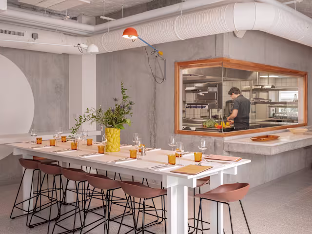Intérieur de restaurant moderne avec une table à manger blanche entourée de tabourets marron, des murs en béton et une grande fenêtre montrant un chef en cuisine. Éclairage chaleureux et arrangement floral coloré dans un vase jaune créent une ambiance contemporaine et accueillante.