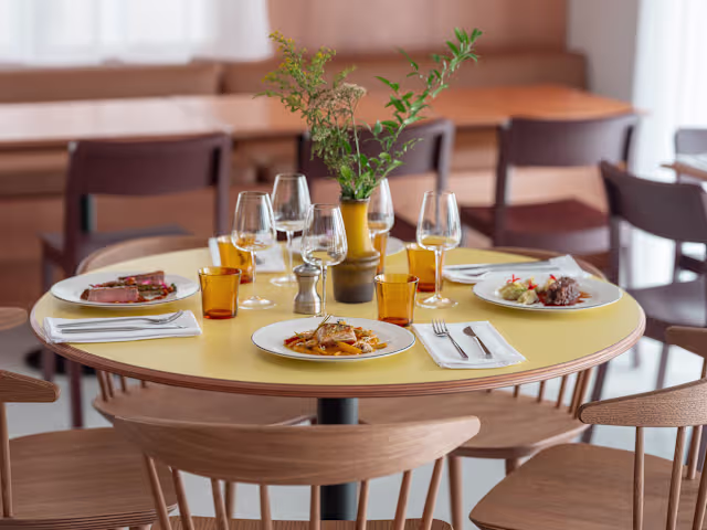 Table ronde avec plusieurs plats colorés, incluant des pâtes garnies d'herbes et d'une sauce légère, ainsi qu'un plat de viande tranchée. Ambiance moderne et accueillante avec un fond en bois chaleureux.