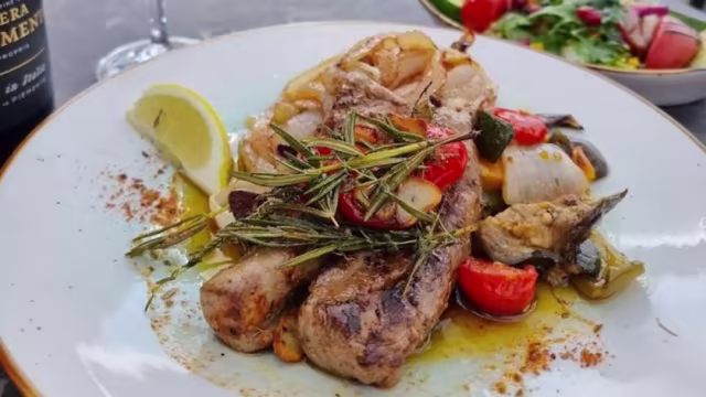 Gegrilltes Fleisch, wahrscheinlich Schwein oder Huhn, garniert mit frischen Kräutern und umgeben von buntem, geröstetem Gemüse wie Kirschtomaten, Paprika und Zwiebeln, serviert mit einer Zitronenscheibe und einem frischen Salat.