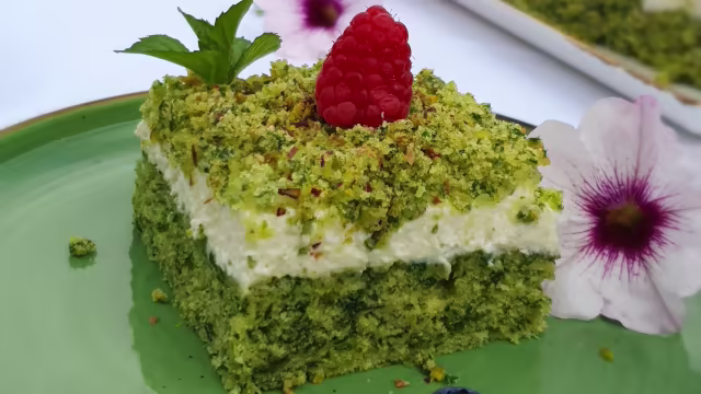 Quadratisches Stück grüner Kuchen mit Pistazien, bedeckt mit cremigem weißen Frosting, garniert mit einer frischen Himbeere und gehackten Pistazien.