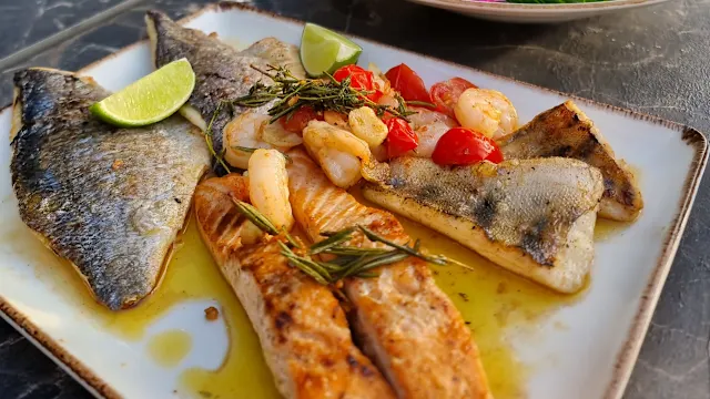 Gegrillte Fischplatte mit zwei Lachsfilets und einem weißen Fisch, garniert mit sautierten Garnelen, gewürfelten Tomaten, Limettenschnitten und Rosmarin, verfeinert mit Olivenöl.