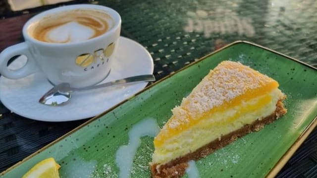 Zitronen-Käsekuchen mit cremiger Füllung und knusprigem, goldbraunem Boden, bestäubt mit Puderzucker, serviert mit einer Tasse Kaffee mit Latte-Art.