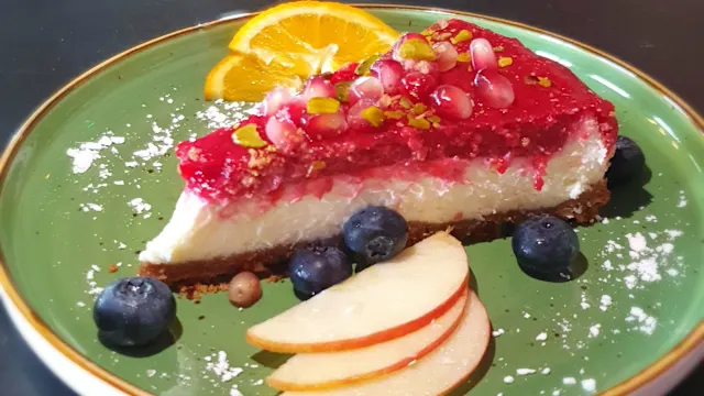 Käsekuchen mit cremiger Füllung, überzogen mit roter Fruchtkompott, auf einem krümeligen Graham-Cracker-Boden, garniert mit Granatapfelkernen und gehackten Pistazien sowie frischen Blaubeeren und dünn geschnittenem Apfel.