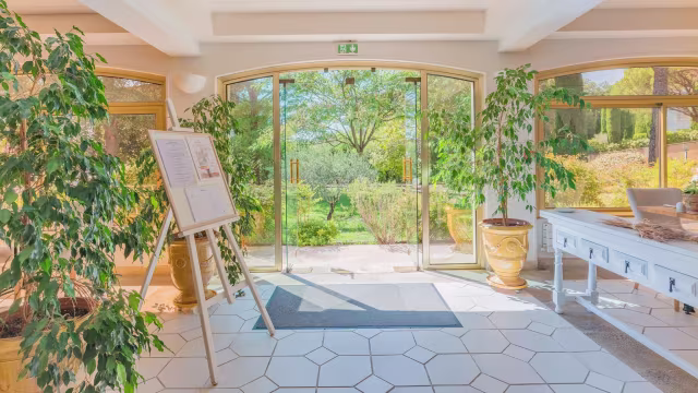 Entrée de restaurant lumineuse avec de grandes portes en verre, décorée de plantes en pot et de carreaux hexagonaux blancs, créant une ambiance accueillante et naturelle.