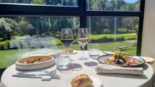 Table élégamment dressée avec deux plats distincts : à gauche, un assortiment de viandes fines, probablement séchées, garni de jeunes pousses et de fleurs comestibles ; à droite, un poisson grillé, probablement du saumon, accompagné de légumes colorés comme des radis et des asperges, le tout rehaussé d'une sauce légère. Deux verres de vin rouge et un verre d'eau complètent cette expérience culinaire raffinée.