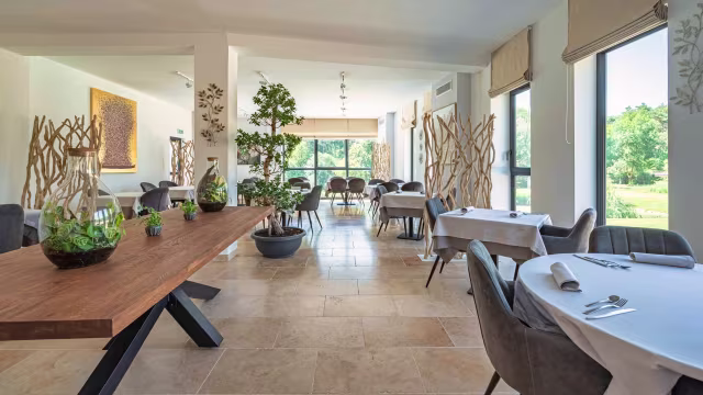 Intérieur moderne et aéré avec de grandes fenêtres, chaises grises élégantes autour de nappes blanches, et une ambiance sereine propice à un repas détendu.