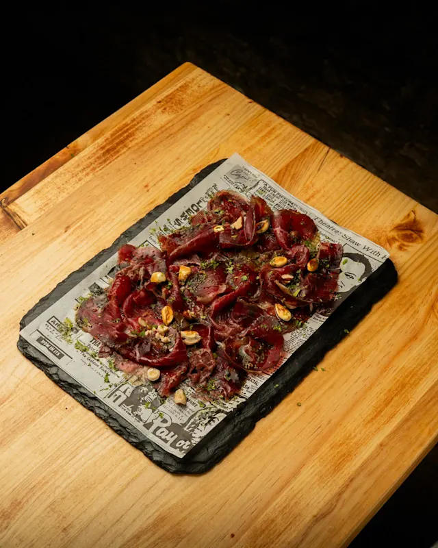 CECINA ADEREZADA Y CANCHITA FRITA Disfruta de un exquisito jamón de ternera, finamente laminado y marinado con una vibrante vinagreta de cilantro y avellanas que realza su sabor. Acompañado de crujiente canchita frita, que añade un toque de textura y unos chorritos de lima fresca.