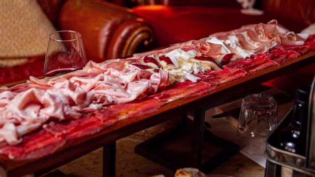Tagliere di salumi con prosciutto, salame e burrata, accompagnato da pane fresco e un bicchiere di vino rosso.