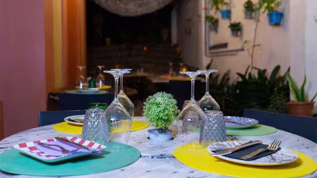 Ambiente acogedor de restaurante al aire libre con mesa redonda decorada con manteles coloridos y plantas colgantes.