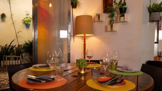Interior acogedor de un restaurante con mesa de madera, platos coloridos y copas de vino, iluminado suavemente por una lámpara.