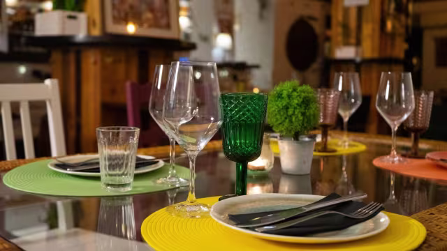 Mesa de comedor elegantemente dispuesta con manteles de colores, cristalería sofisticada y un toque de vegetación.