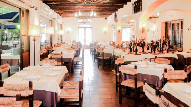 Interior espacioso con vigas de madera y luz cálida, mesas con manteles blancos y sillas rústicas, decorado con esculturas y arte, creando un ambiente acogedor y elegante.