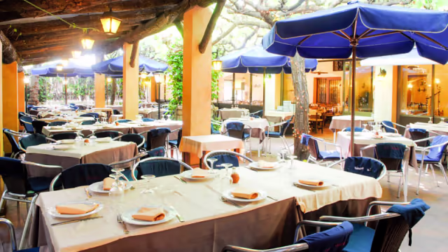 Área de comedor al aire libre con mesas bajo sombrillas azules, rodeada de plantas verdes, ideal para disfrutar de comidas al fresco.
