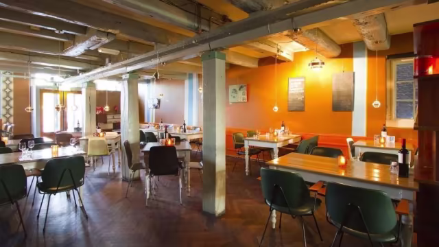 Het interieur van het restaurant heeft een warme en uitnodigende sfeer met houten tafels en moderne stoelen. De muren zijn in een rijke oranje tint geschilderd, met zichtbare balken en industriële verlichting, wat bijdraagt aan een gezellige eetomgeving.