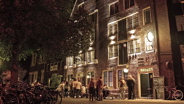 Levendige restaurantgevel bij nacht met warme verlichting, grote ramen en een drukke sfeer, omringd door fietsen en een gevoel van gemeenschap.