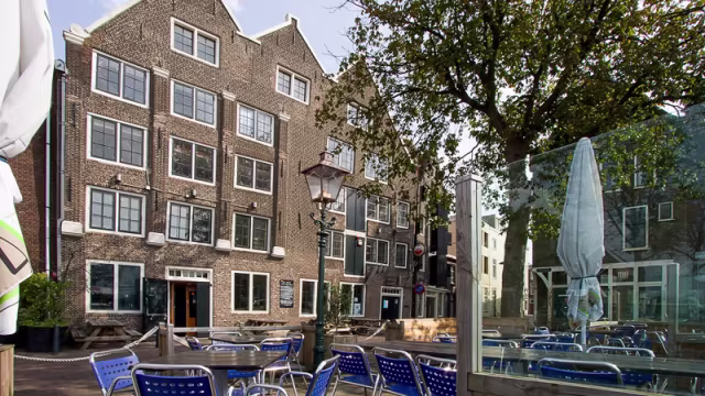 Buitenterras van een restaurant met blauwe metalen stoelen rond houten tafels, omringd door traditionele bakstenen gebouwen en een grote boom die schaduw biedt, ideaal voor dineren in de buitenlucht.