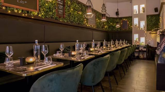 Het restaurantinterieur heeft een modern en uitnodigend ontwerp met weelderige groenvoorzieningen aan de muren, wat een frisse en natuurlijke sfeer creëert. De zitplaatsen bestaan uit luxe, donkergroene stoelen rond strakke tafels, verlicht door kaarsen en decoratieve lantaarns.