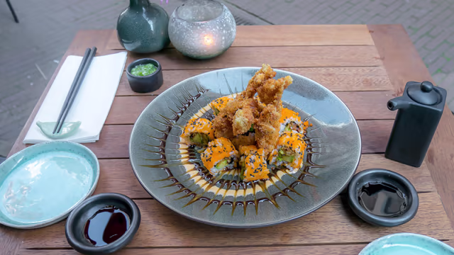 Sushi plateau met kleurrijke sushirollen gevuld met verse vis of groenten, getopt met zwarte sesamzaadjes en krokante tempura, geserveerd met een donkere saus en kleine schaaltjes sojasaus en wasabi.