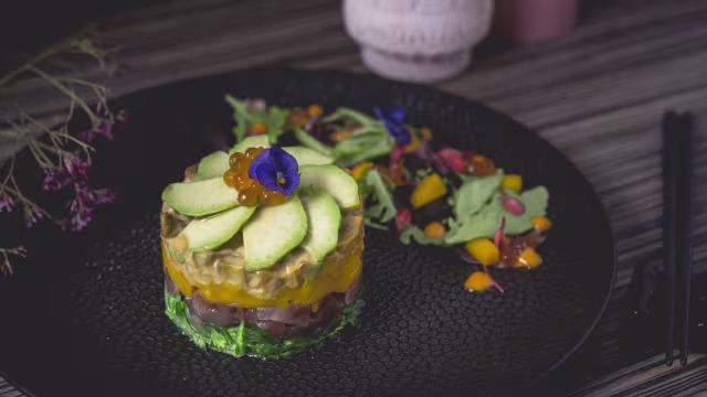 Gelaagde schotel met fijngehakte groenten, waaronder sla en kruiden, gevolgd door blokjes paprika en komkommer, en afgewerkt met gesneden avocado. Versierd met viskuit en een eetbare bloem, geserveerd met een bijgerecht van gemengde sla en kleurrijke fruitblokjes in vinaigrette.